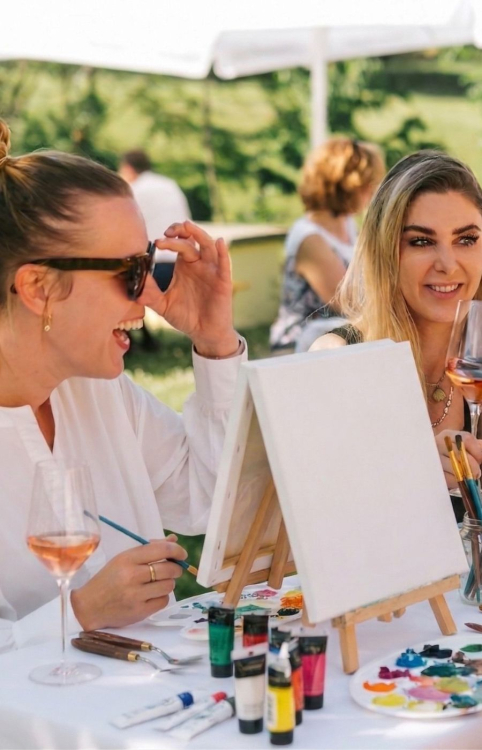 Zwei Damen sitzen vor einem Keilrahmen, malen ein Bild und trinken Rosé