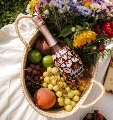 In einem Picknickkorb mit Obst und Blumen liegt eine Spring Edition, auf der Picknickdecke daneben liegt Käse und Brot