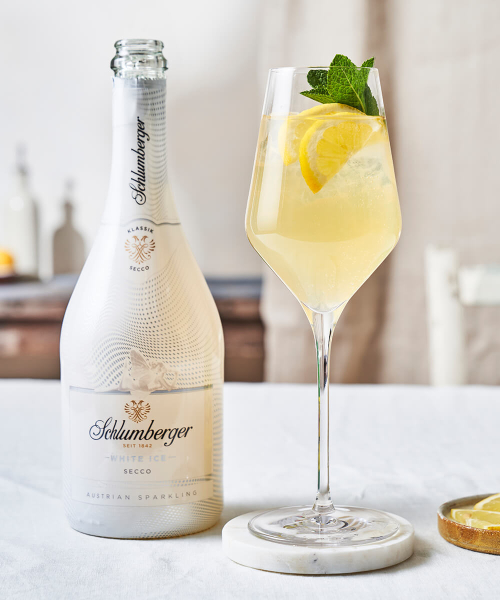 Flasche Schlumberger White Ice Secco neben einem Weinglas mit einem Limonada Spritz Cocktail, dekoriert mit Zitronenscheiben und frischer Minze, auf einem hellen Tisch arrangiert.