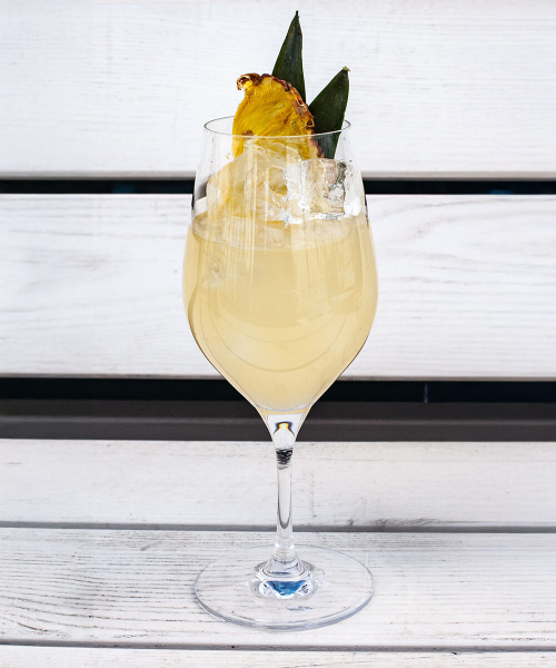 Ein Weinglas gefüllt mit einem Mai Tai Spritz Cocktail und einer Ananasscheibe als Garnitur, auf einer weißen Bank