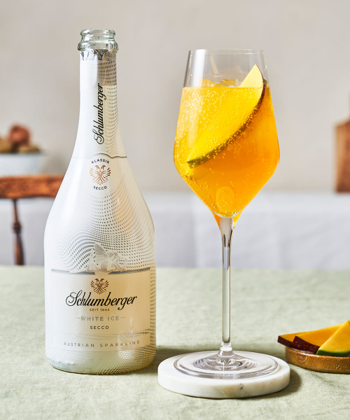 Flasche Schlumberger White Ice Secco neben einem Weinglas mit Mango Spritz, dekoriert mit einer Mangoscheibe, auf einem hellen Tisch mit einer Schale Mango-Stückchen.