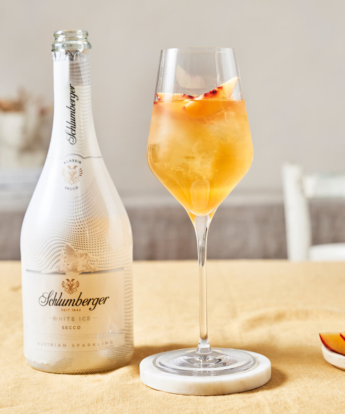 Flasche Schlumberger White Ice Secco neben einem Weinglas mit einem Metaxa Pfirsich Spritz Cocktail, dekoriert mit einer Pfirsichspalte, auf einem hellen Tisch arrangiert.