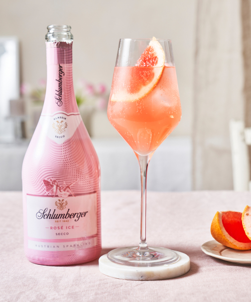 Flasche Schlumberger Rosé Ice Secco neben einem Weinglas mit Pink Spritz, dekoriert mit einer Grapefruitscheibe, auf einem hellen Tisch mit Grapefruit-Stückchen.