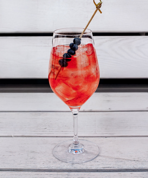 Ein Weinglas gefüllt mit einem Pornstar Spritz Cocktail und einem Heidelbeer-Spieß auf einer weißen Bank