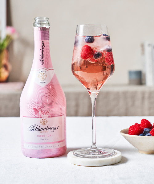 Flasche Schlumberger Rosé Ice Secco neben einem Weinglas mit einem Purple Berry Spritz Cocktail, dekoriert mit frischen Beeren, auf einem hellen Tisch arrangiert.