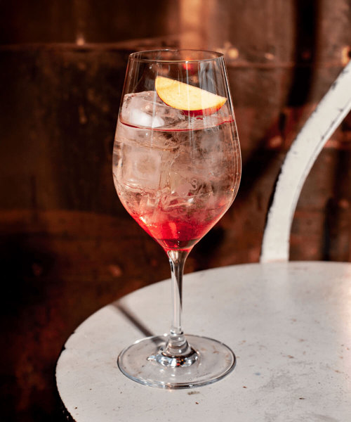 Ein Weinglas gefüllt mit einem Sex on the Spritz Cocktail und einer Pfirsichspalte als Garnitur, auf einem runden Tisch in einem Kellergewölbe