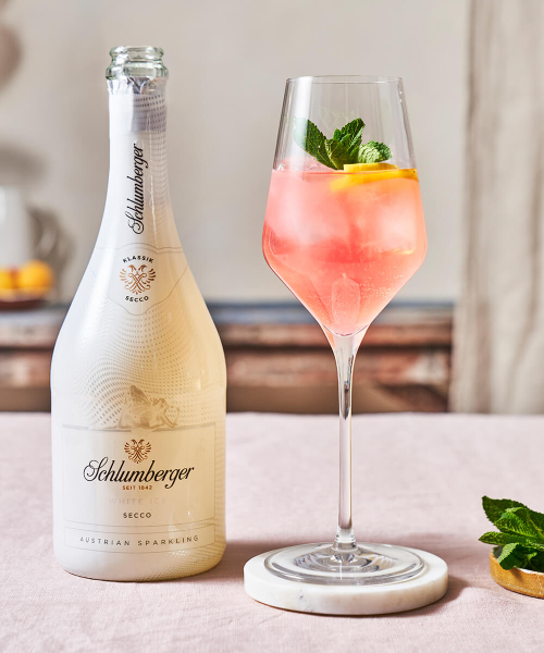 Flasche Schlumberger White Ice Secco neben einem Weinglas mit einem White Melon Spritz Cocktail, dekoriert mit Zitronenscheiben und Minze, auf einem hellen Tisch arrangiert.
