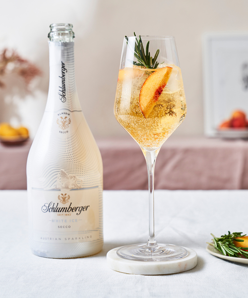 Flasche Schlumberger White Ice Secco neben einem Weinglas mit einem White Peach SpritzCocktail, dekoriert mit einer Pfirsichscheibe und einem Rosmarinzweig, auf einem hellen Tisch arrangiert.