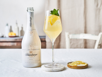 Flasche Schlumberger White Ice Secco neben einem Weinglas mit einem Limonada Spritz Cocktail, dekoriert mit Zitronenscheiben und frischer Minze, auf einem hellen Tisch arrangiert.