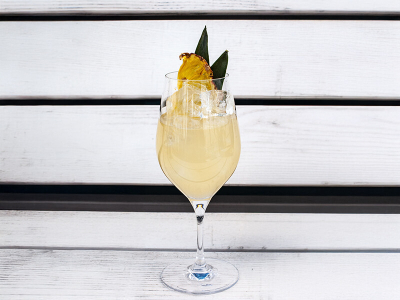 Ein Weinglas gefüllt mit einem Mai Tai Spritz Cocktail und einer Ananasscheibe als Garnitur, auf einer weißen Bank