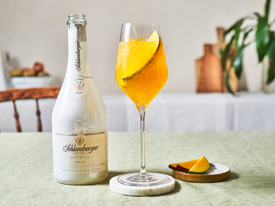 Flasche Schlumberger White Ice Secco neben einem Weinglas mit Mango Spritz, dekoriert mit einer Mangoscheibe, auf einem hellen Tisch mit einer Schale Mango-Stückchen.