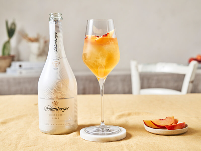 Flasche Schlumberger White Ice Secco neben einem Weinglas mit einem Metaxa Pfirsich Spritz Cocktail, dekoriert mit einer Pfirsichspalte, auf einem hellen Tisch arrangiert.