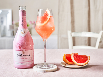 Flasche Schlumberger Rosé Ice Secco neben einem Weinglas mit Pink Spritz, dekoriert mit einer Grapefruitscheibe, auf einem hellen Tisch mit Grapefruit-Stückchen.