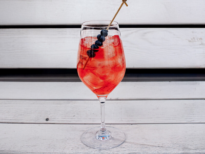 Ein Weinglas gefüllt mit einem Pornstar Spritz Cocktail und einem Heidelbeer-Spieß auf einer weißen Bank