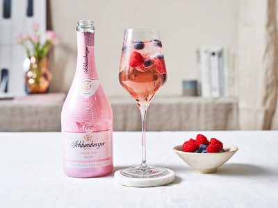 Flasche Schlumberger Rosé Ice Secco neben einem Weinglas mit einem Purple Berry Spritz Cocktail, dekoriert mit frischen Beeren, auf einem hellen Tisch arrangiert.