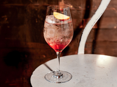 Ein Weinglas gefüllt mit einem Sex on the Spritz Cocktail und einer Pfirsichspalte als Garnitur, auf einem runden Tisch in einem Kellergewölbe