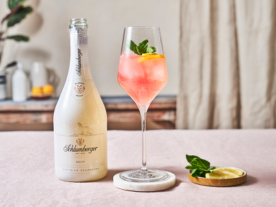 Flasche Schlumberger White Ice Secco neben einem Weinglas mit einem White Melon Spritz Cocktail, dekoriert mit Zitronenscheiben und Minze, auf einem hellen Tisch arrangiert.