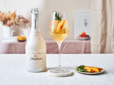 Flasche Schlumberger White Ice Secco neben einem Weinglas mit einem White Peach SpritzCocktail, dekoriert mit einer Pfirsichscheibe und einem Rosmarinzweig, auf einem hellen Tisch arrangiert.